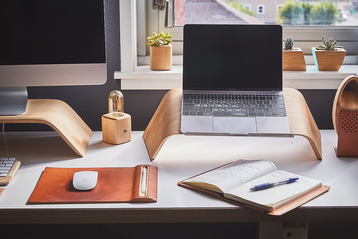 Notebook na stojane, monitor, zápisník a kožená podložka na stole pri okne